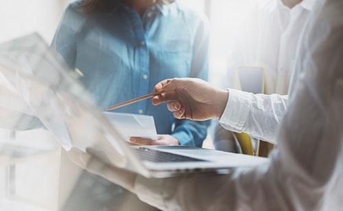 A close-up of a business meeting where professionals are discussing a project. One person in a white shirt is pointing at a laptop screen with a pencil while another holds documents. The scene is illuminated by natural light, emphasizing collaboration and strategic IT consulting.