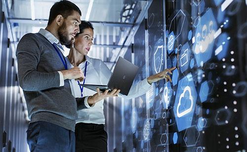 **Alt Text:** Two IT professionals, a man and a woman wearing lanyards, work inside a high-tech server room. The woman is pointing at a futuristic digital interface displaying cloud computing and cybersecurity icons, while the man holds a laptop and observes. The scene highlights advanced technology for IT consulting.