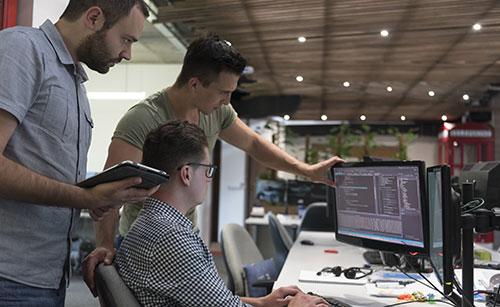 Three software developers collaborate in IT Consulting office space. One person is seated at a dual-monitor setup with code displayed on the screens, while another points at the screen and a third holds a tablet, discussing the project. The workspace has an open-concept design with plants and decorative lighting.